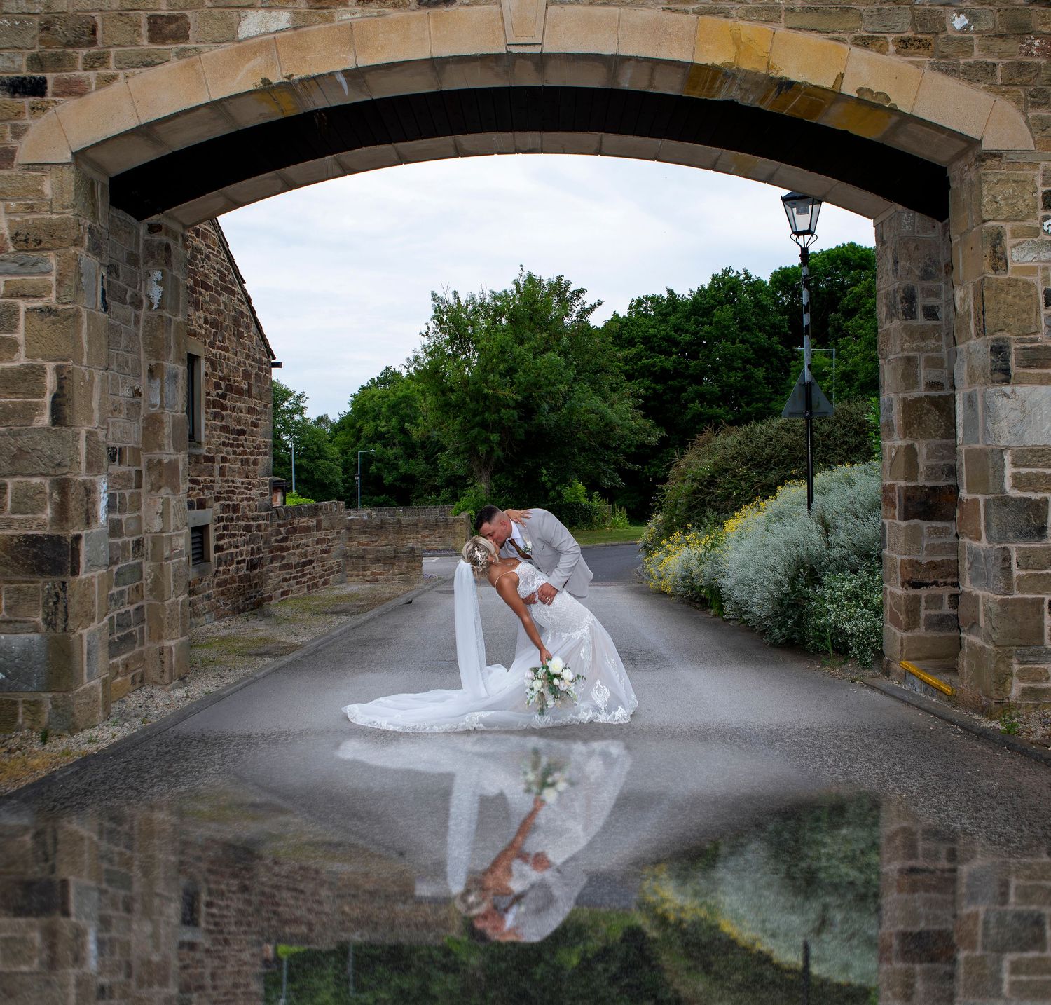 Bride and groom share a romantic kiss under a stone archway, with their reflection visible on the wet pavement below.