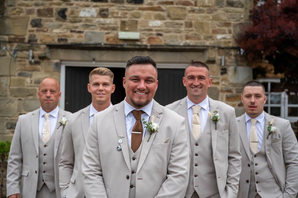 Groom with groomsmen in light grey suits with boutonnieres posing outside stone building.