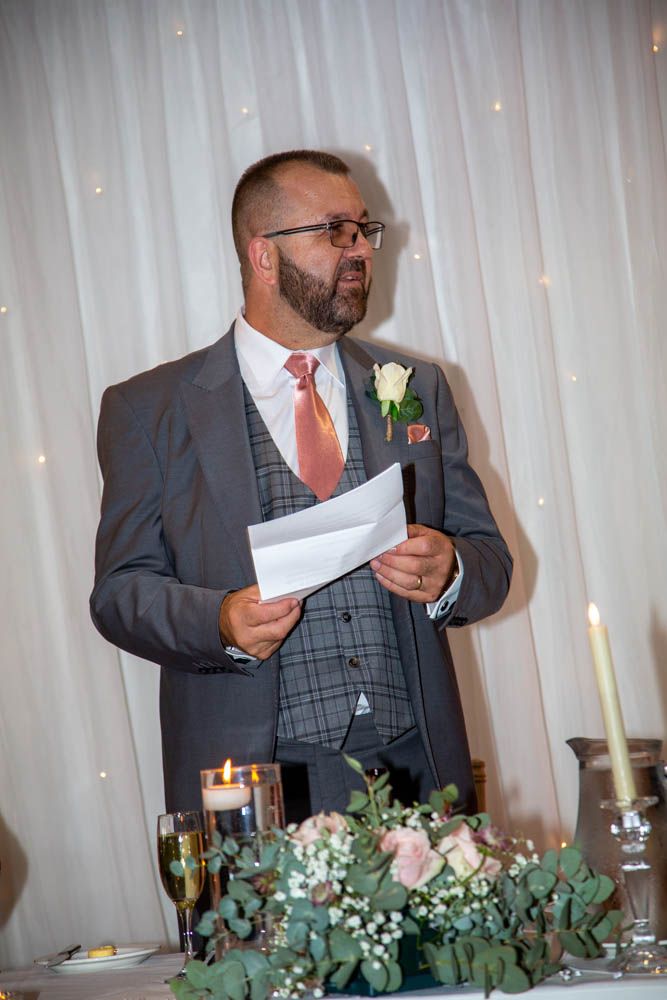 Man in gray suit with pink tie reads speech at wedding reception, with floral arrangements and candles in foreground.