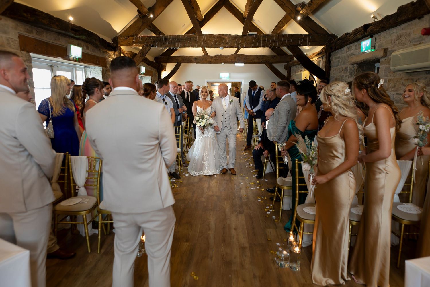 Bride walks down the aisle in a rustic wedding venue at Tankersley Manor with bridesmaids in gold dresses and guests seated on either side.