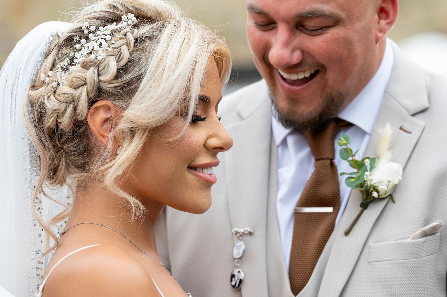 Smiling bride with braided updo and veil beside laughing groom in gray suit with boutonniere on wedding day.