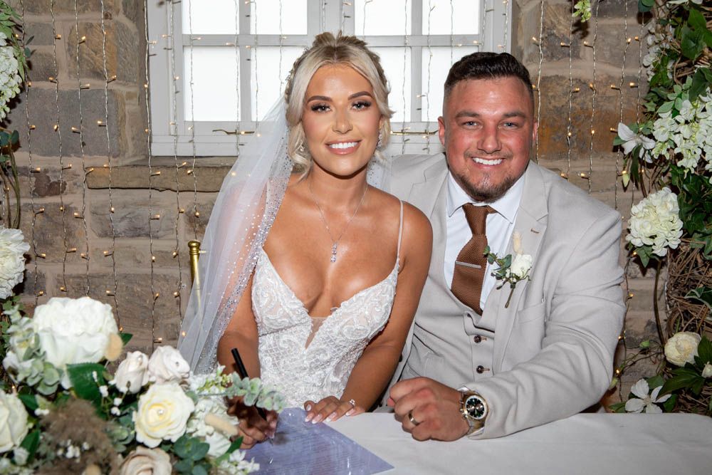 Bride and groom smile at signing table surrounded by white floral arrangements at wedding.