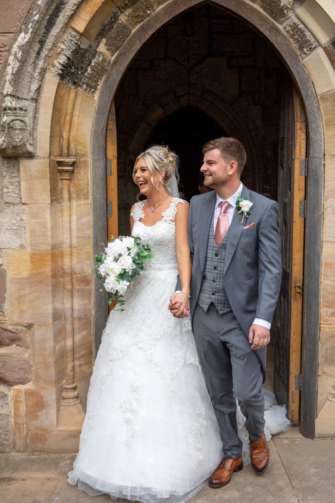 Bride and groom smiling and holding hands outside a stone church doorway, bride holding white bouquet.