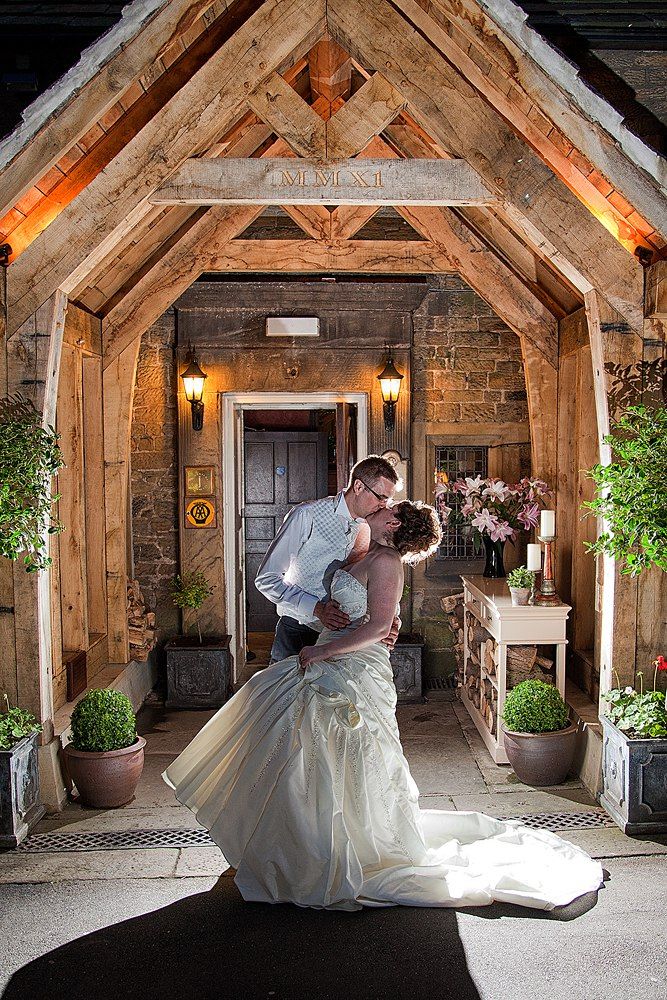 Bride and groom sharing a kiss under a rustic wooden arch at a stone venue.