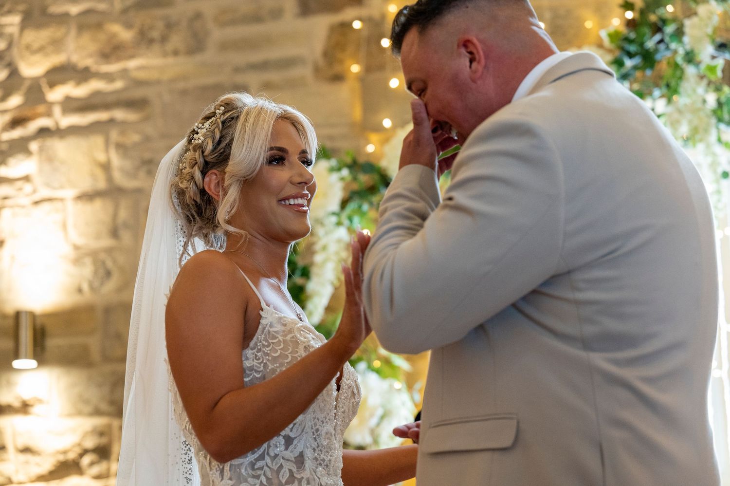 Emotional groom wiping tears as bride smiles during outdoor wedding ceremony with stone wall backdrop.