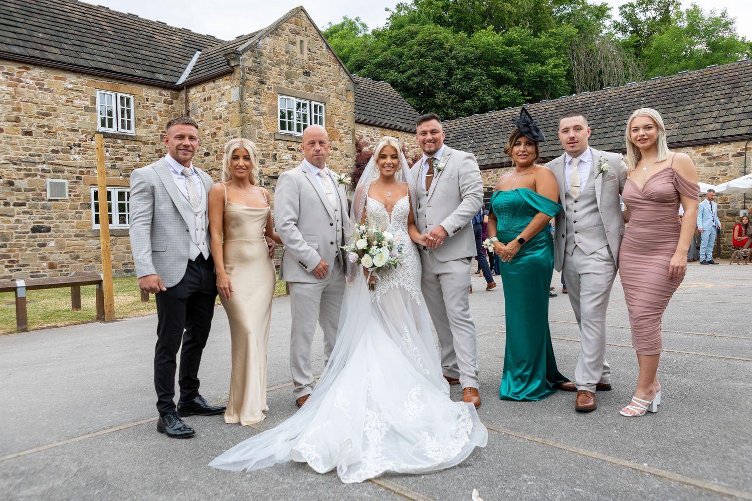 Wedding party poses outside stone venue, bride in white gown with groom and guests in formal attire.