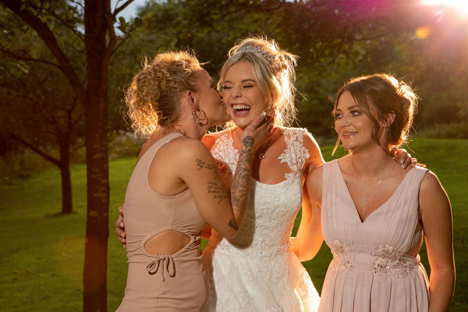 Bride in white lace gown laughing joyfully with two bridesmaids in blush dresses during golden hour outdoor wedding.