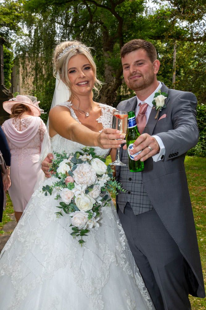 Bride and groom toasting with champagne and beer on their wedding day, bride holding white floral bouquet.