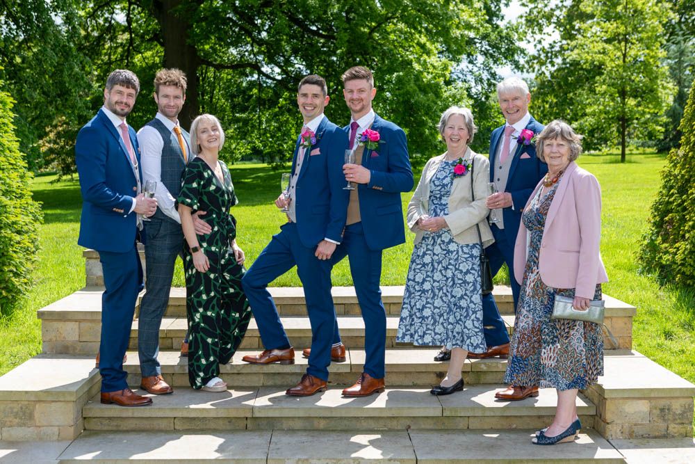A smiling wedding group of seven stands on stone steps outdoors in a lush green garden setting of Rudding Park Hotel.
