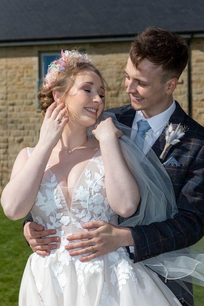 A smiling bride and groom embrace outdoors on their wedding day, she in lace gown with floral crown, he in tartan suit.