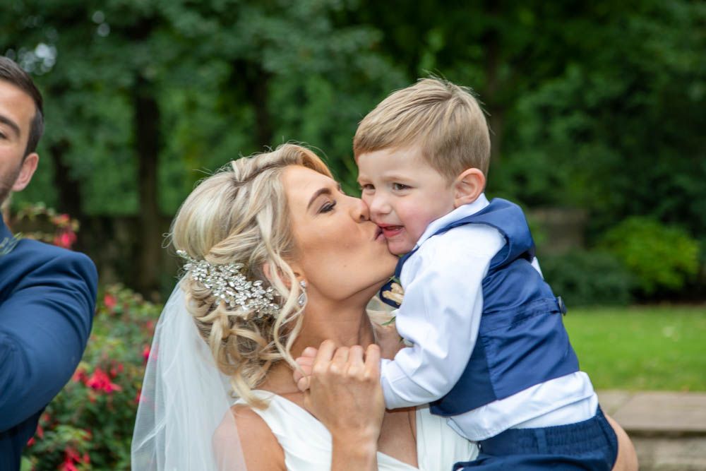 Bride in white wedding gown gives a kiss to a young boy in a navy suit.