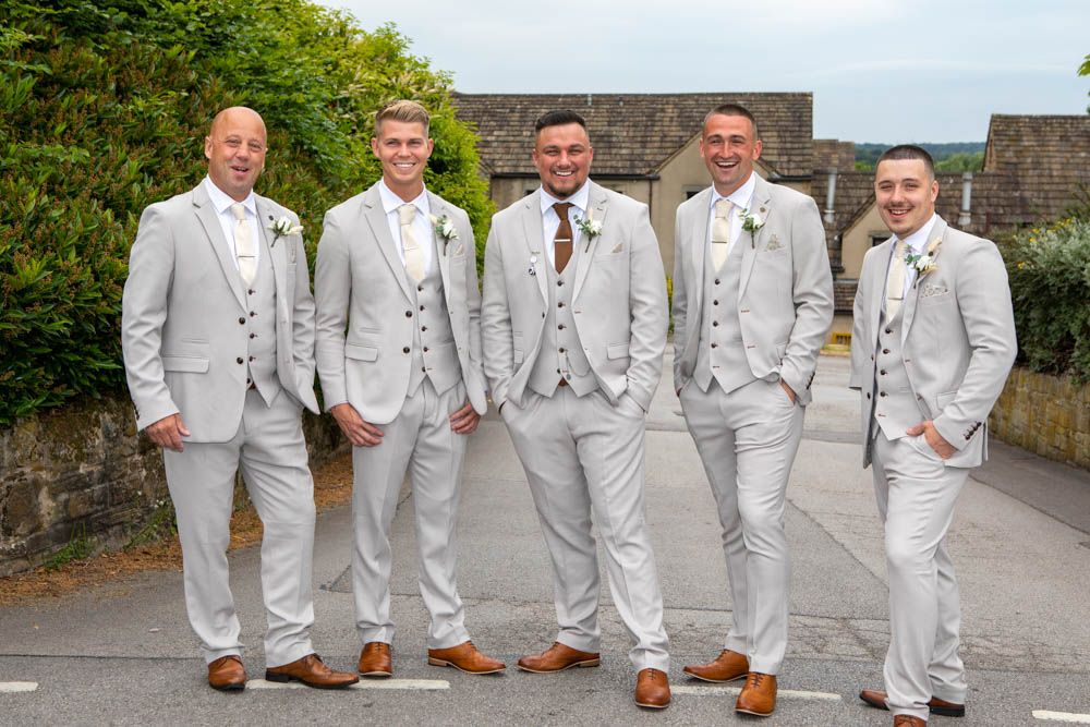 Five groomsmen in matching light grey suits and brown shoes pose outdoors on wedding day.