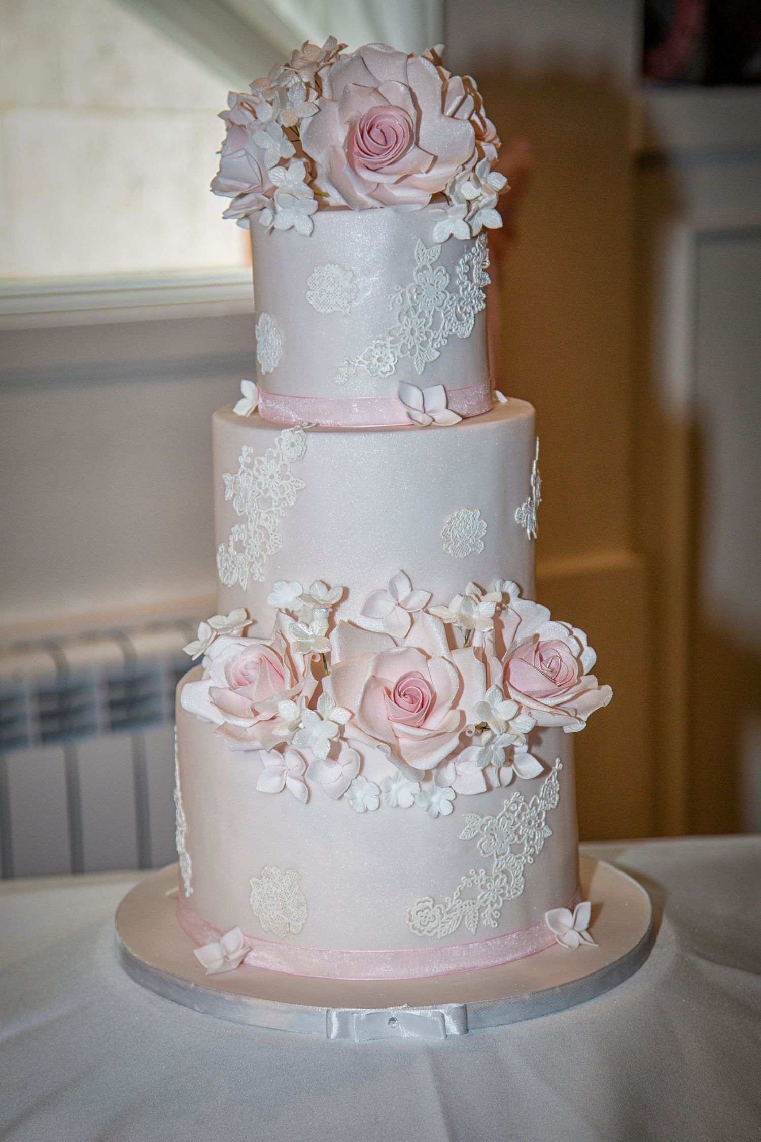 Elegant four-tier white wedding cake with pink sugar roses and white lace fondant details on a silver base.