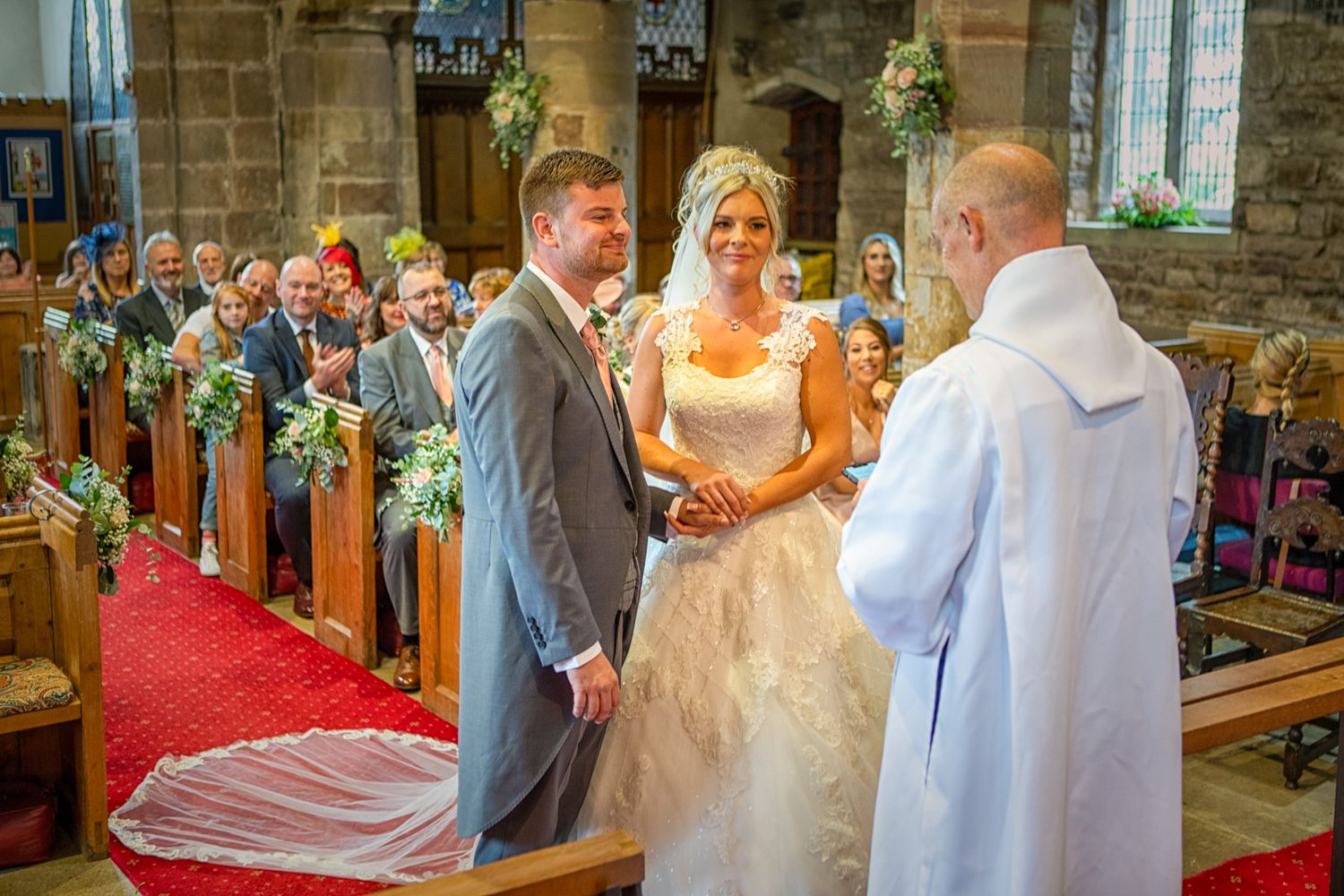 Bride and groom exchange vows at a traditional stone church - All Saint's, wedding ceremony with bridesmaids and guests seated in pews.