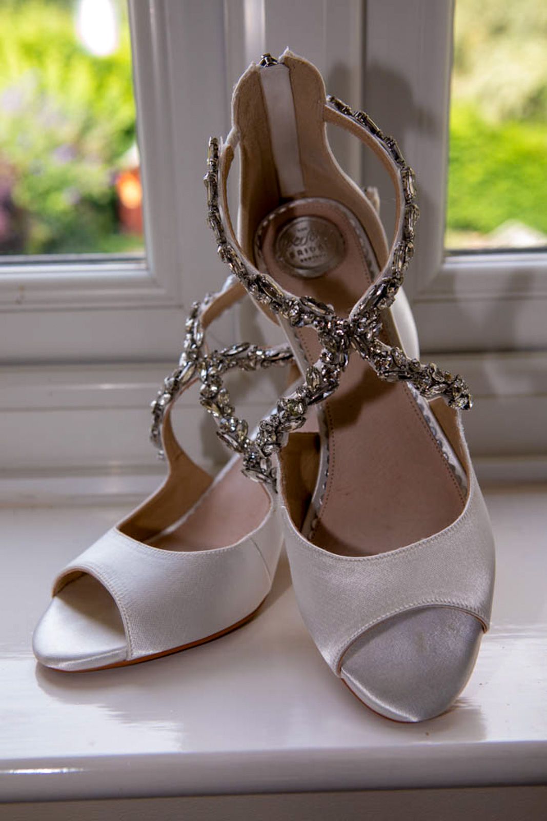 Elegant bridal heels with crystal embellished straps displayed on white windowsill.