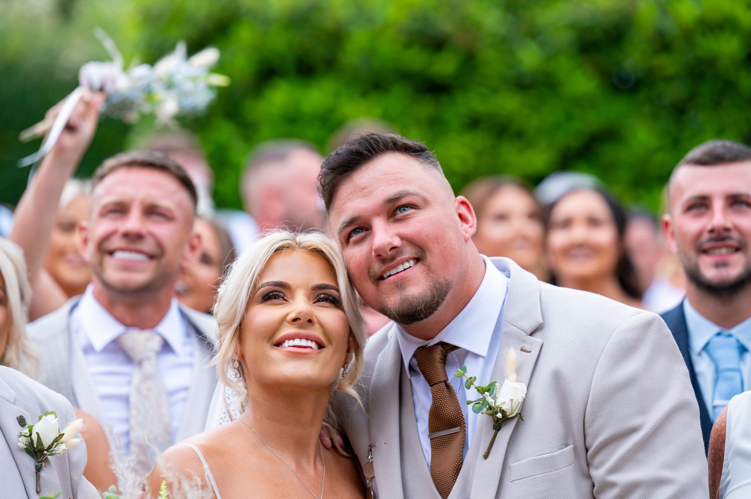 Happy newlyweds smiling joyfully at outdoor wedding ceremony with guests celebrating in background.