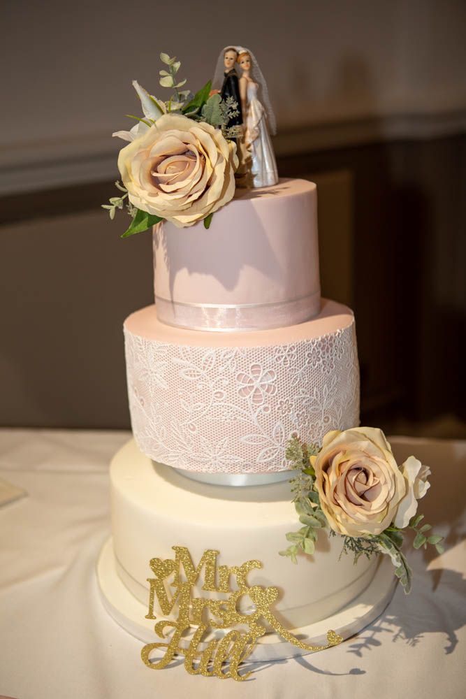 Three-tier wedding cake with lace detail, roses, and Mrs & Mrs Hill gold topper.