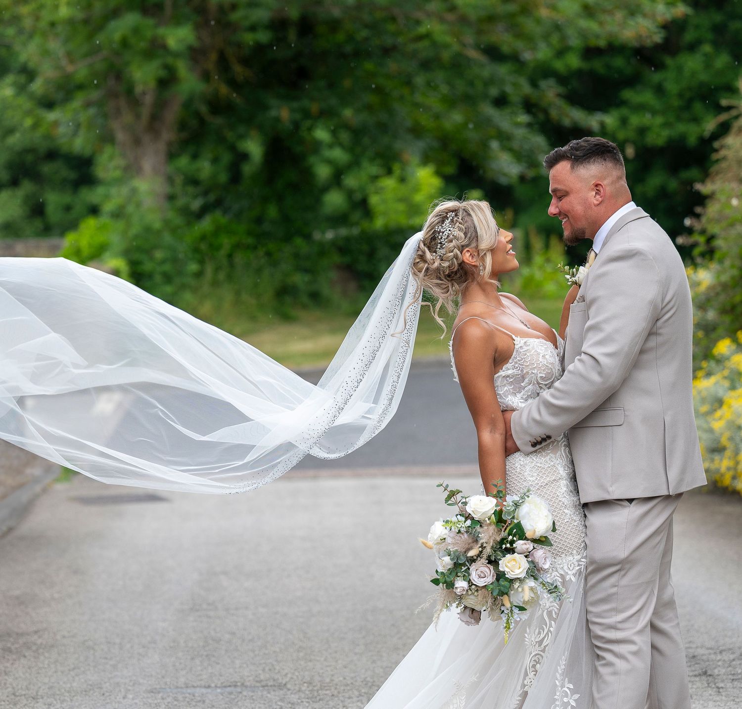 Bride and groom share romantic moment outdoors, her white veil flowing in wind, holding bouquet, groom in gray suit.