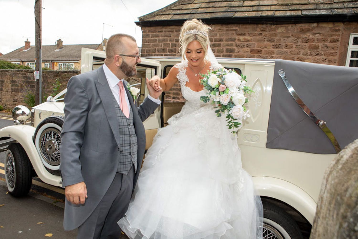Bride in white gown holding bouquet assisted by groom in gray suit beside vintage cream wedding car.