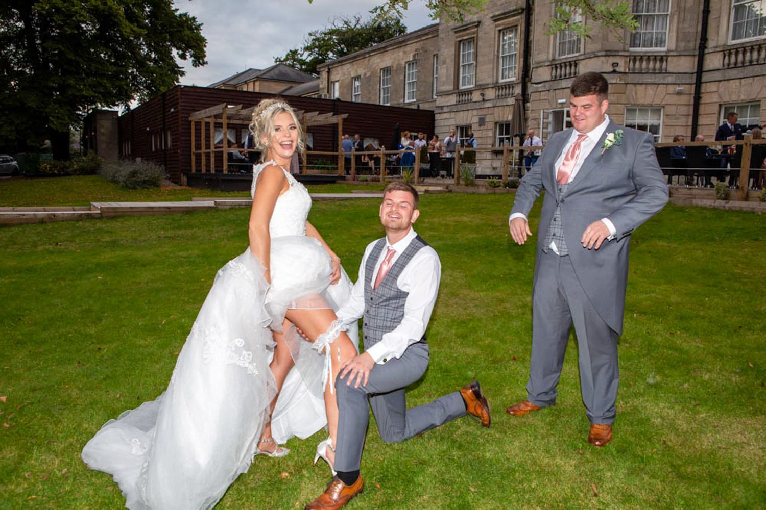 Bride and groom pose playfully with groomsman on green lawn outside elegant stone wedding venue - Aston Hall Hotel.