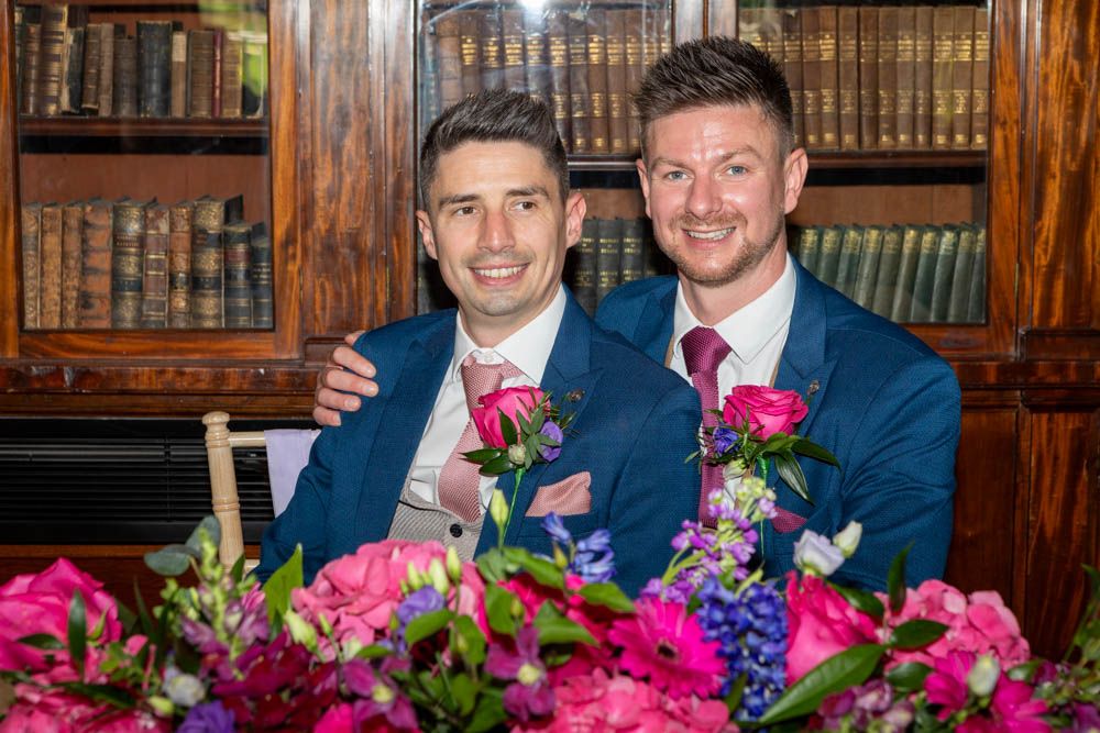 Two grooms in blue suits smile at wedding table with pink floral arrangements in front of a bookcase.