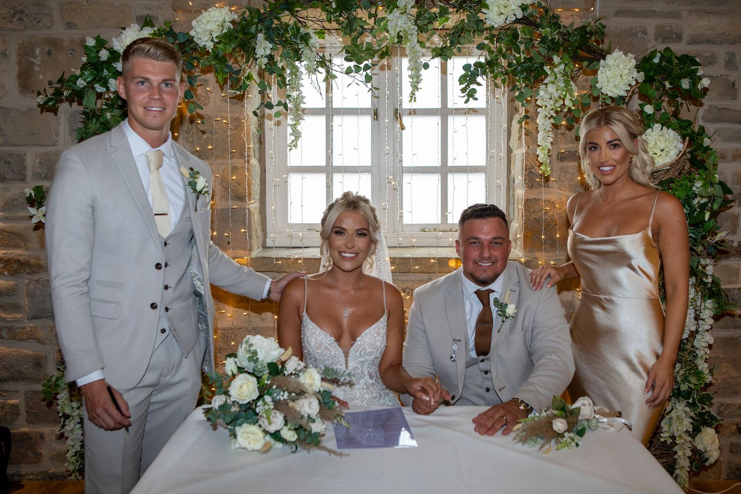 Bride and groom signing wedding register at decorated table with two guests standing beside them in elegant venue.