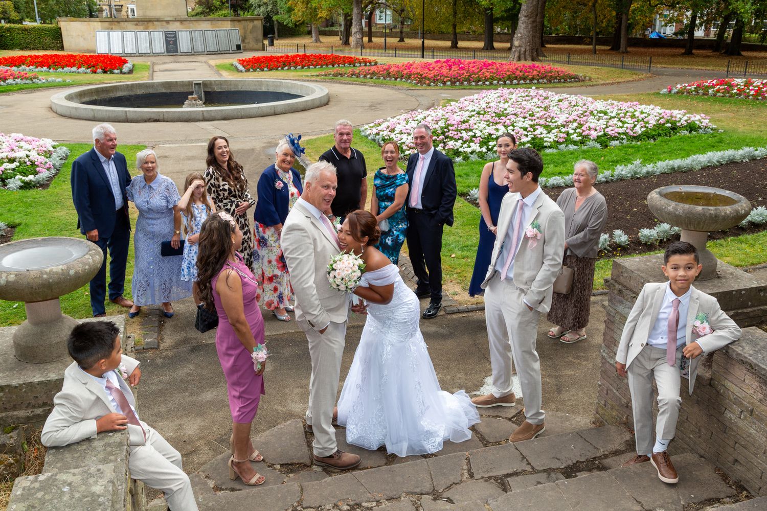 Outdoor wedding ceremony in a garden with bride and groom surrounded by guests near a fountain with colorful flowers.