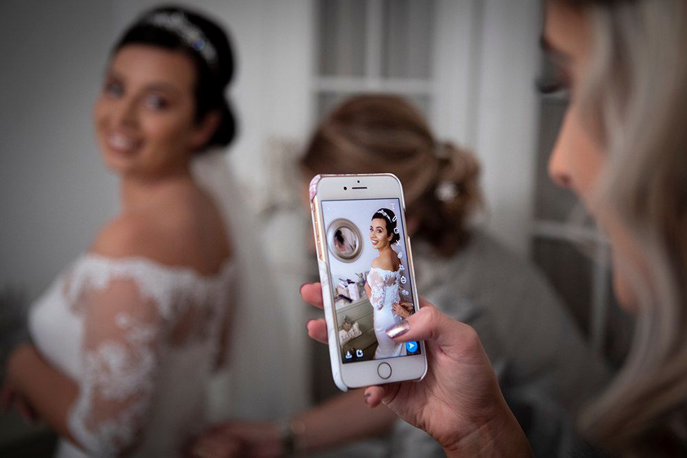 Wedding guest photographs smiling bride in lace gown using smartphone at bridal preparation.