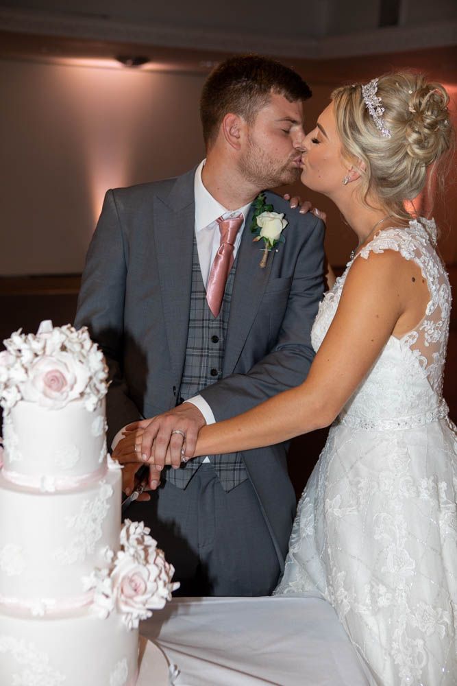 Bride and groom share a kiss while cutting their white wedding cake decorated with pink roses.