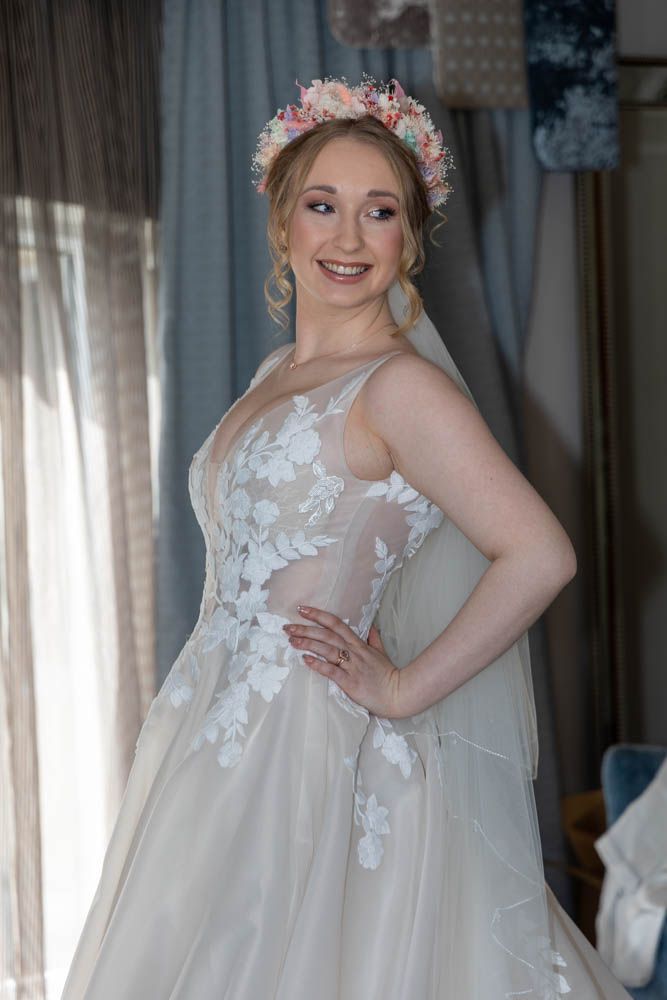 Smiling bride in floral lace wedding gown and pink flower crown poses elegantly indoors.