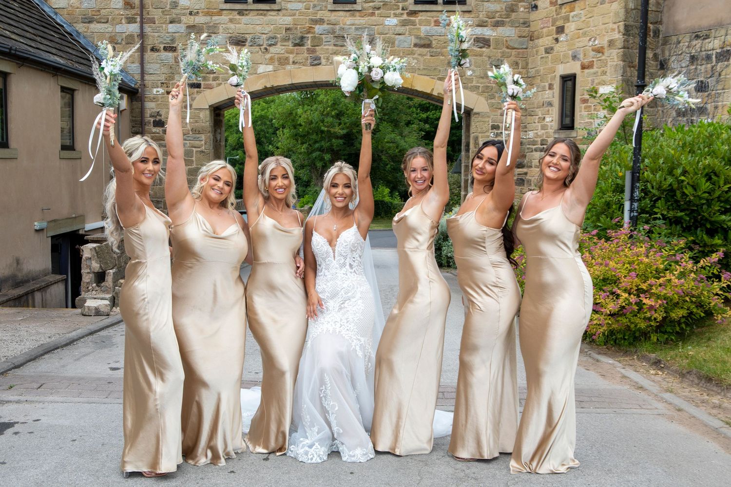 Bride in white lace gown with six bridesmaids in gold satin dresses raising bouquets outside stone venue.
