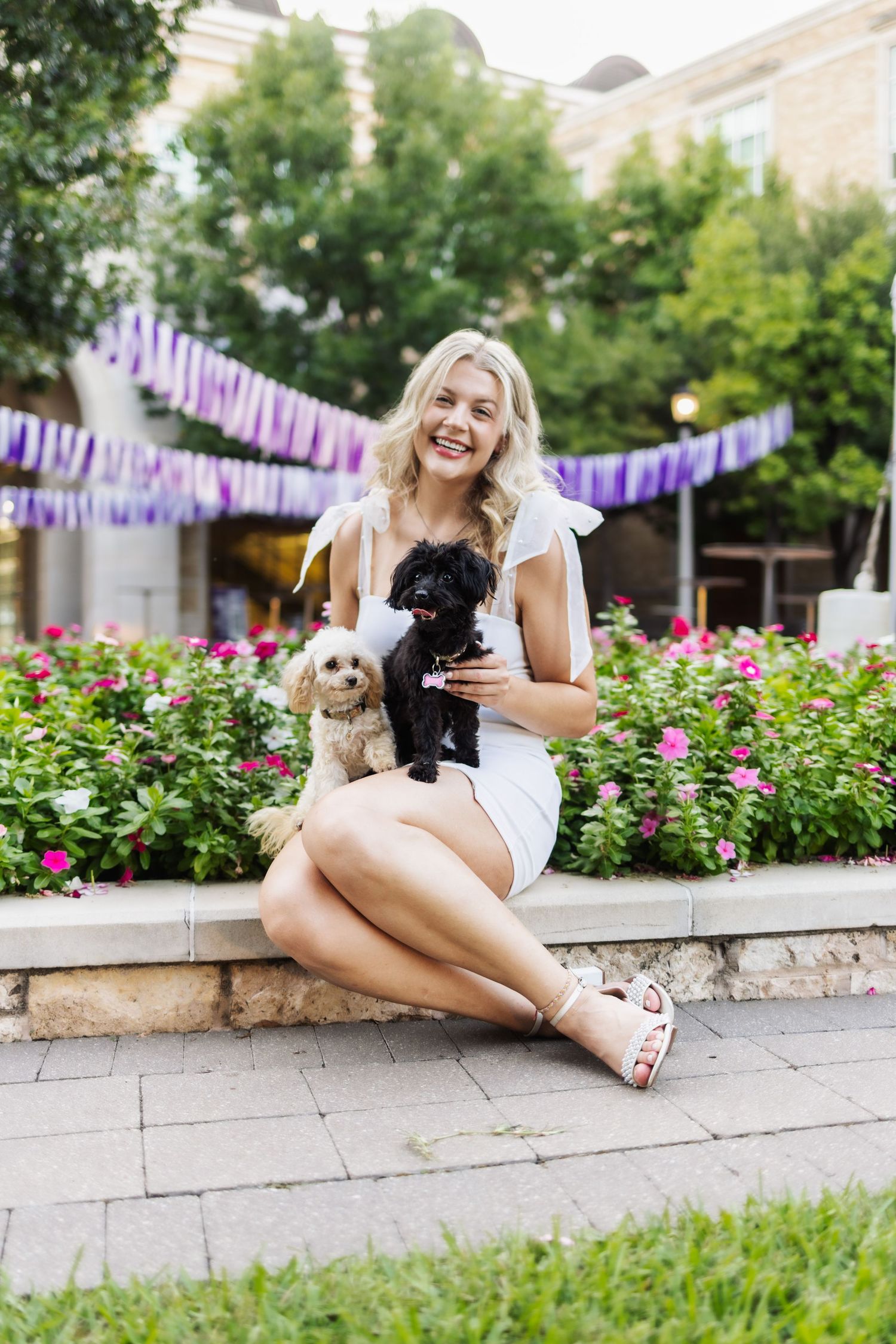 Smiling blonde woman in white dress sitting outdoors holding two small dogs near flowers.
