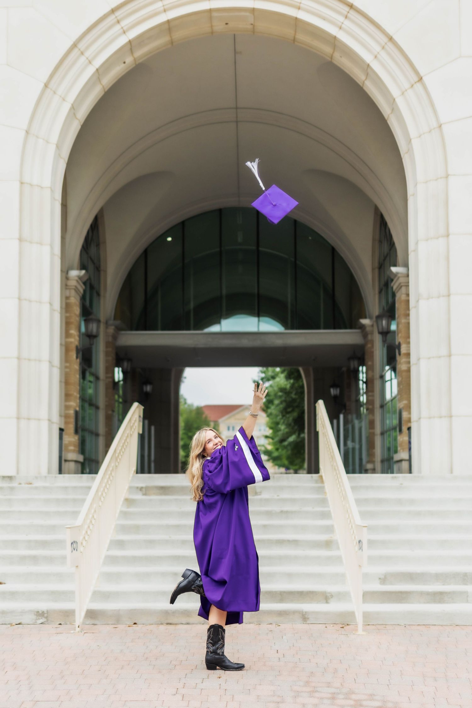 Graduate in purple gown tosses cap in air before grand arched building entrance.