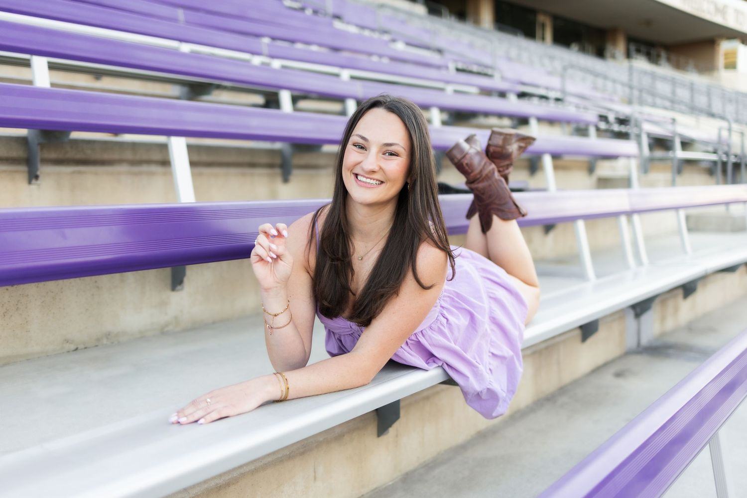 Young woman in purple outfit smiling and posing on purple stadium bleachers in a cheerful outdoor setting.