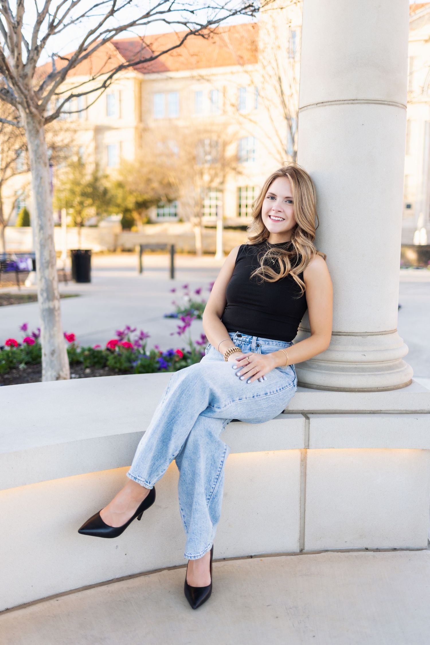 Young woman in black top and light jeans sitting on stone ledge by column, smiling outdoors.