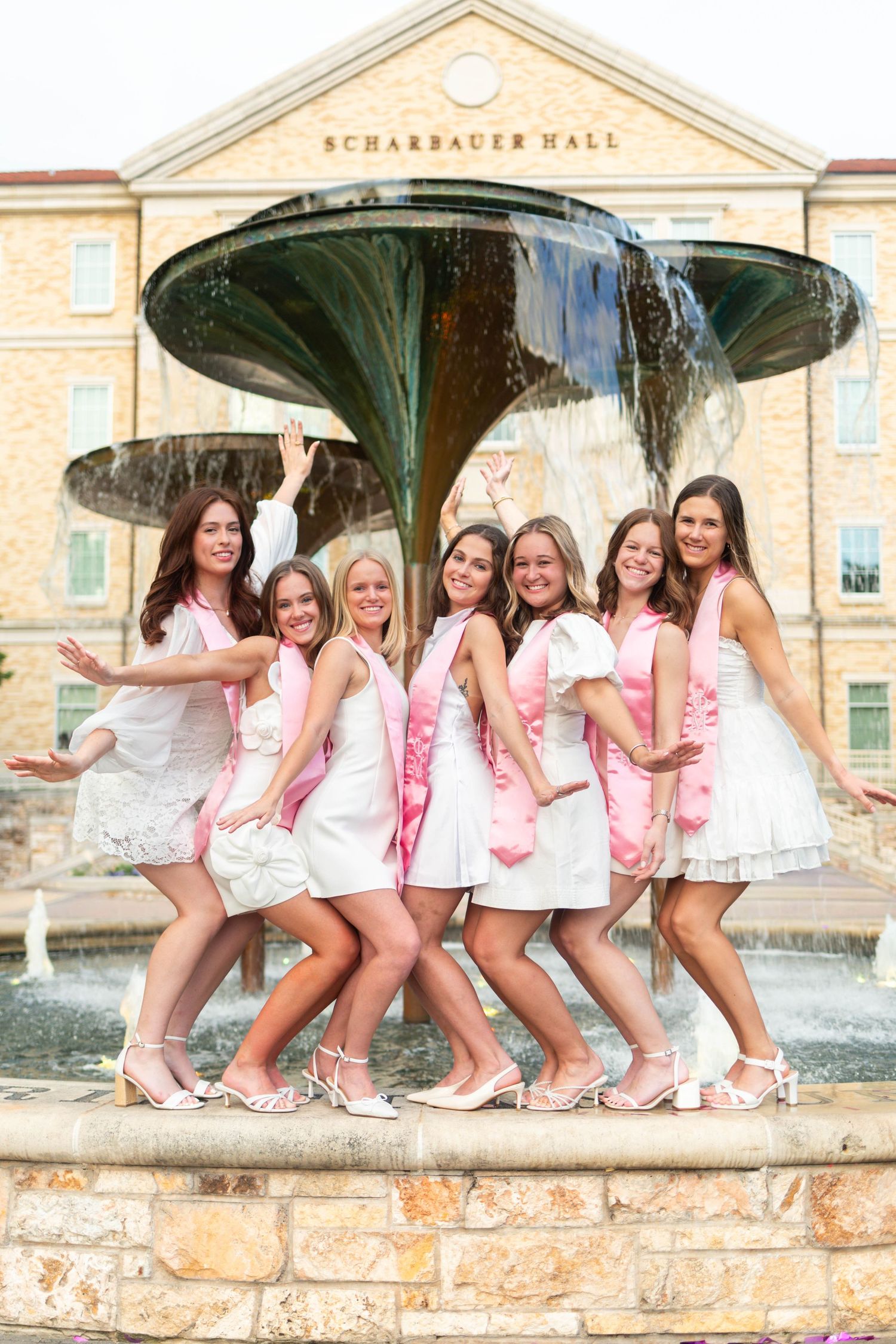 Seven young women in white dresses and pink sashes pose together by a fountain outside Scharbauer Hall.