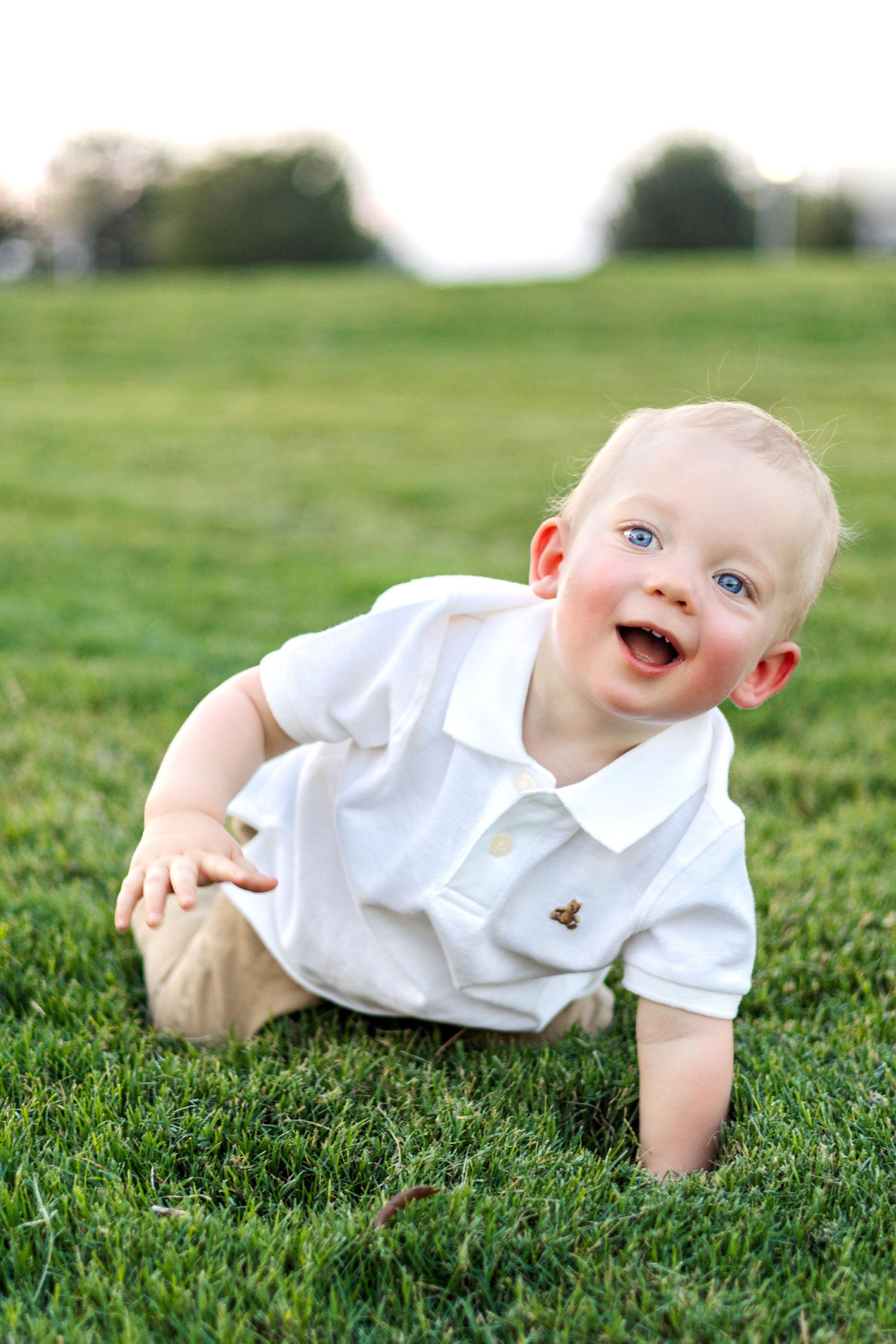 Happy baby boy with blue eyes crawling on green grass, wearing a white polo shirt and khaki pants outdoors.