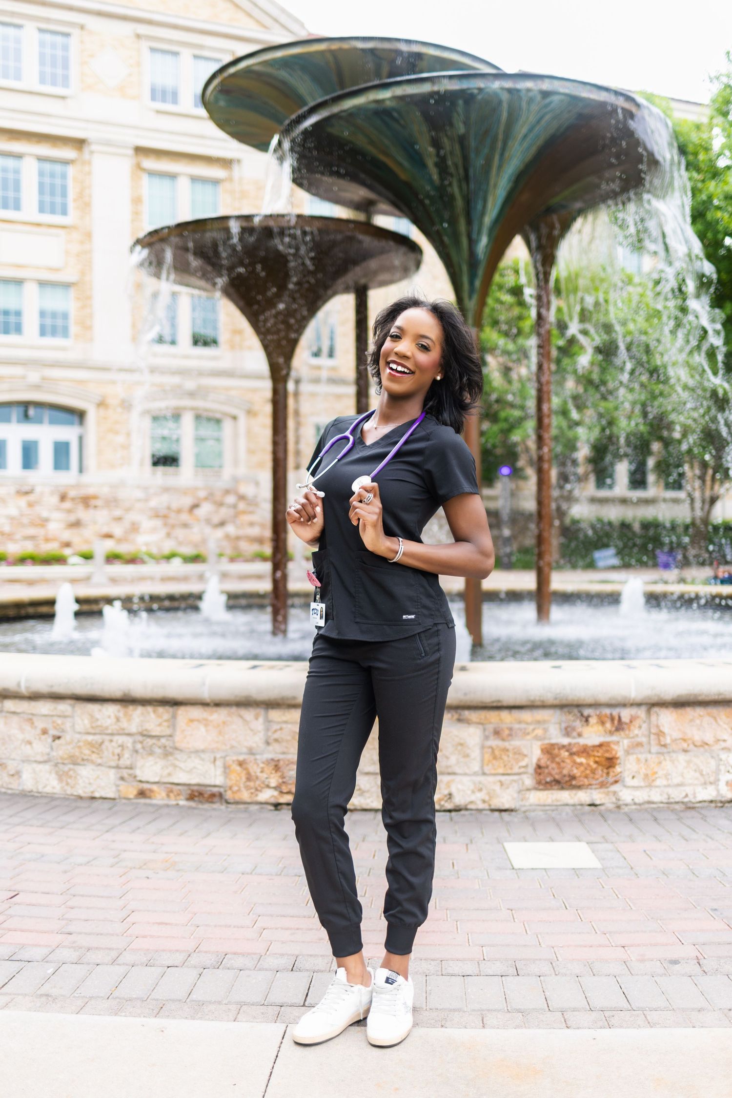 Smiling TCU nursing graduate in black scrubs poses in front of a campus fountain with stethoscope.