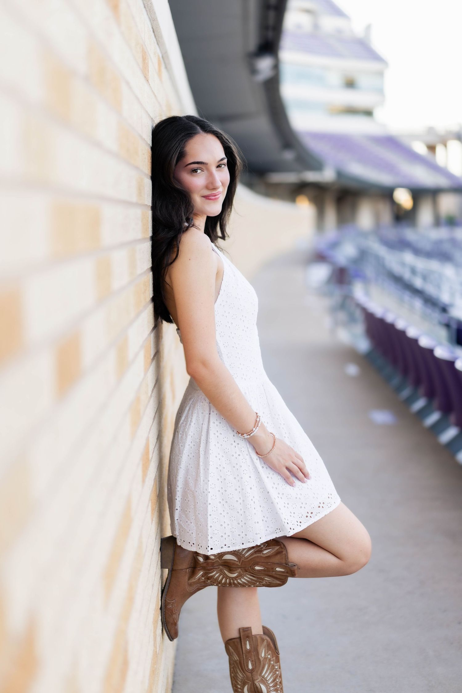 Young woman in white dress and cowboy boots leaning against a stadium wall, smiling confidently.