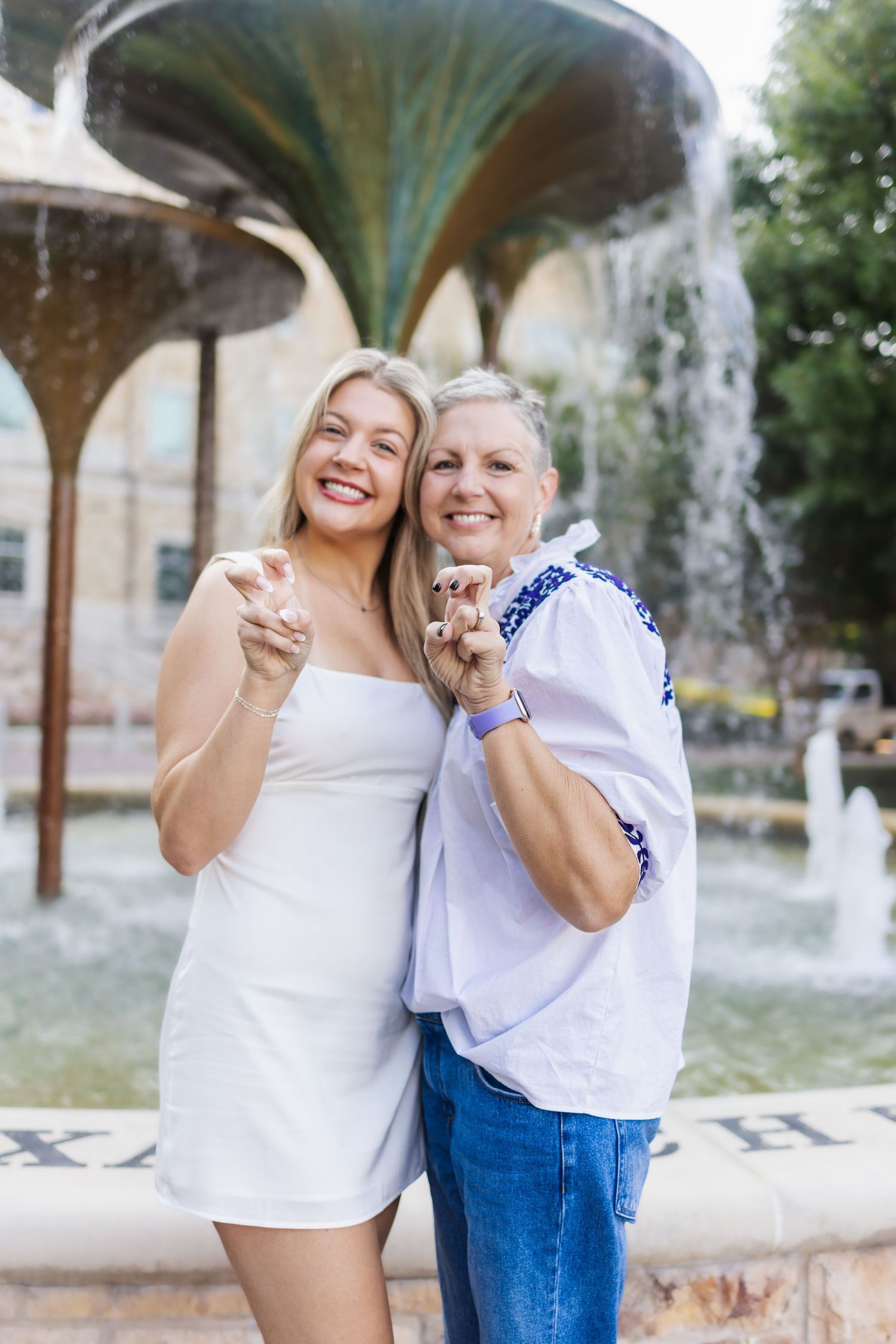 Two smiling women pose together in front of a decorative campus fountain at a graduation celebration.