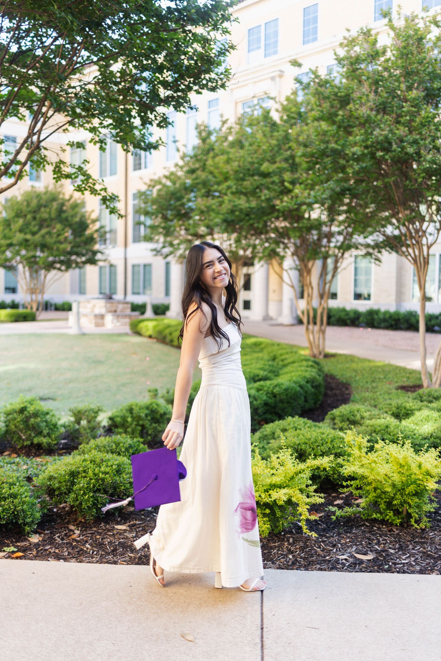 Smiling TCU graduate in white dress holds purple cap while walking through a lush campus garden.