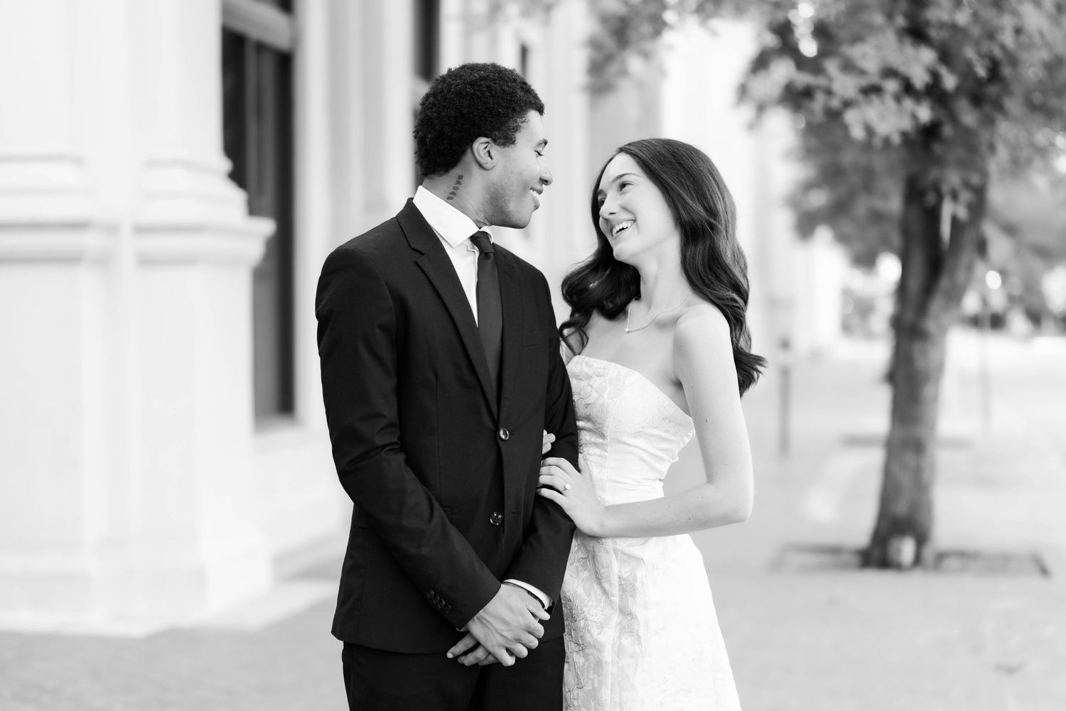 Black and white photo of a smiling couple in wedding attire standing outdoors, groom in suit and bride in strapless gown.
