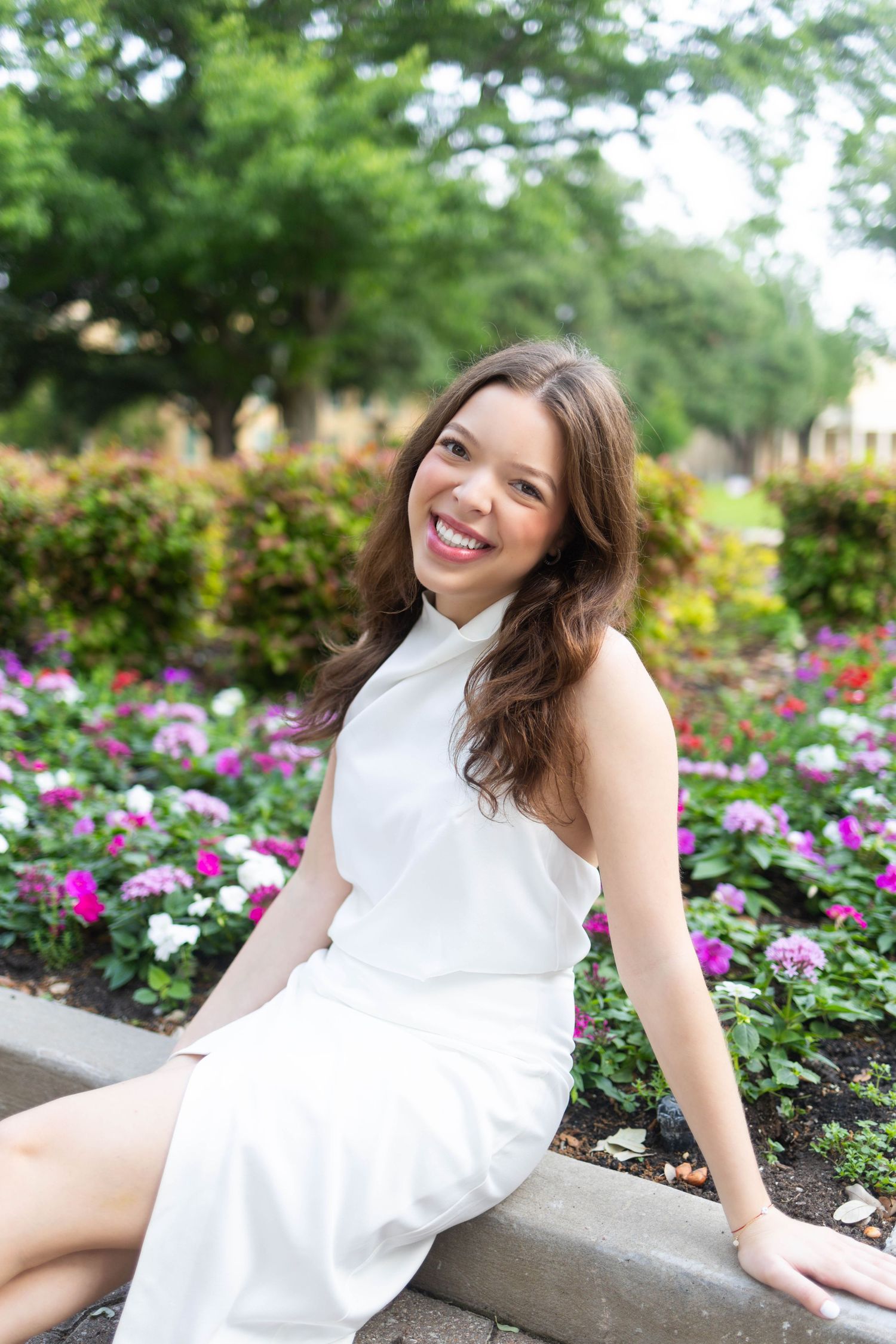 Young woman in white dress smiling in a colorful flower garden with lush green trees in the background.