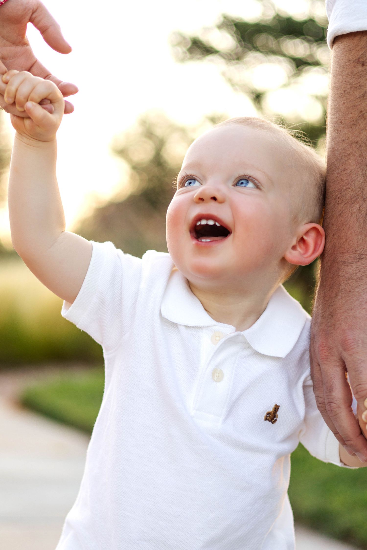 Happy blue-eyed baby boy in white polo shirt laughing while holding adult hand outdoors in golden sunlight.