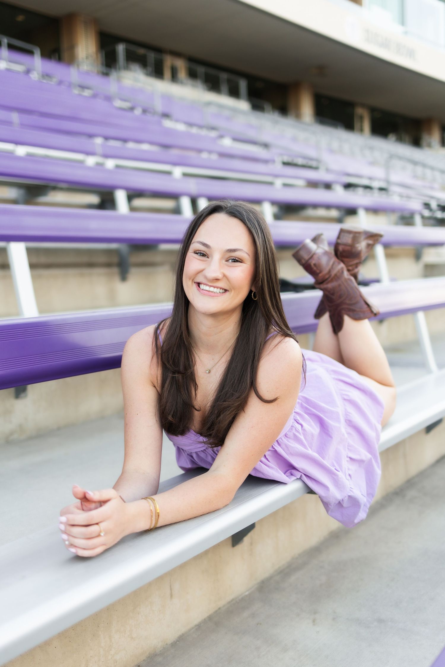Smiling brunette woman in lavender dress lies on purple stadium bleachers wearing brown boots.