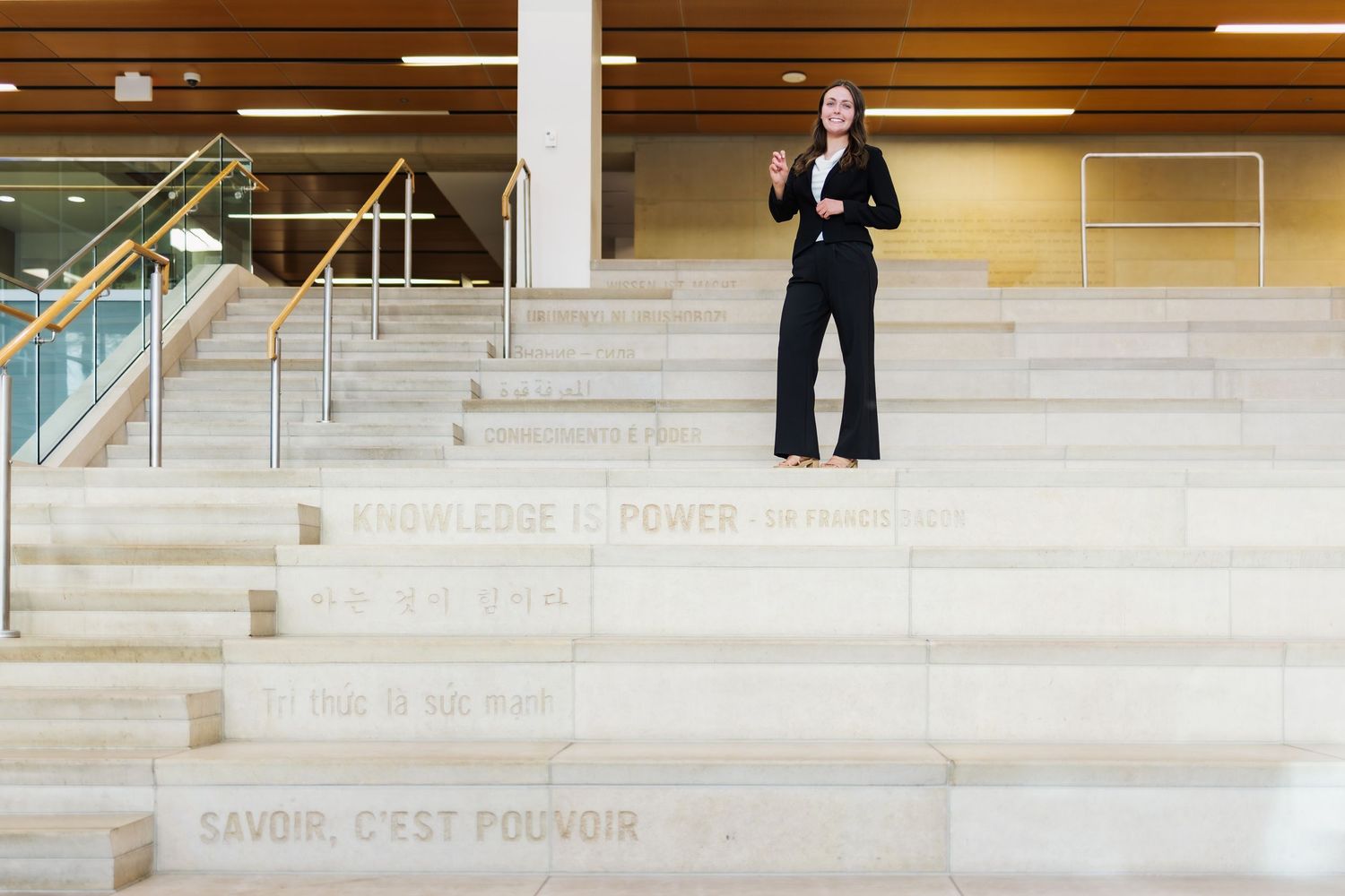 Woman in black suit stands on marble stairs with 'Knowledge is Power' inscription in modern building.