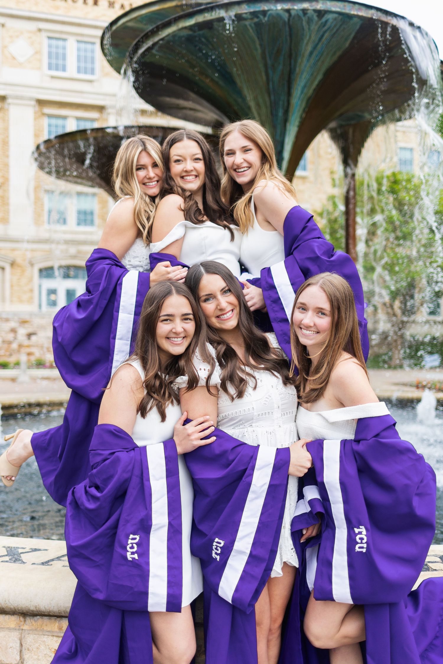Six young women in purple TCU graduation robes pose together smiling in front of a fountain.