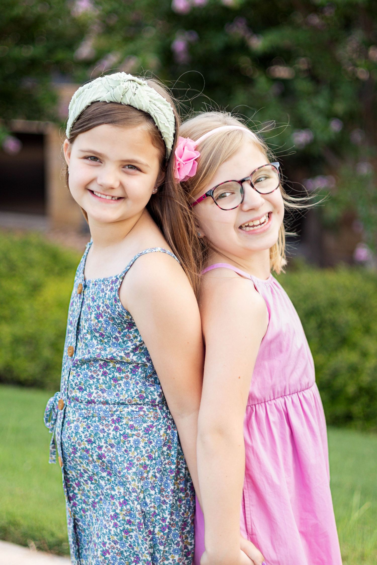 Two smiling girls in summer dresses pose back-to-back outdoors, one wearing glasses and a pink flower hair clip.