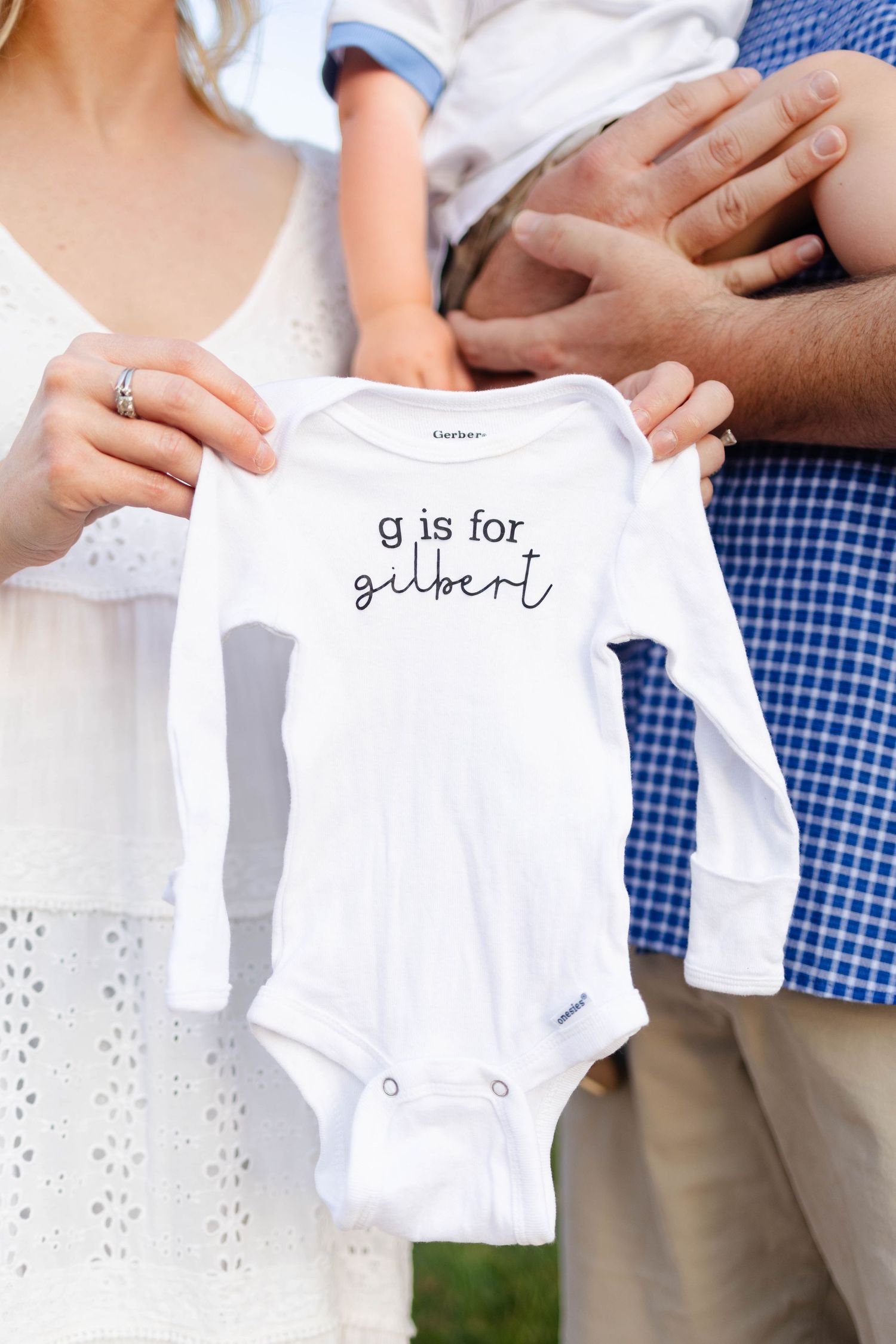 Parents holding a white Gerber onesie that reads g is for gilbert, pregnancy announcement photo.