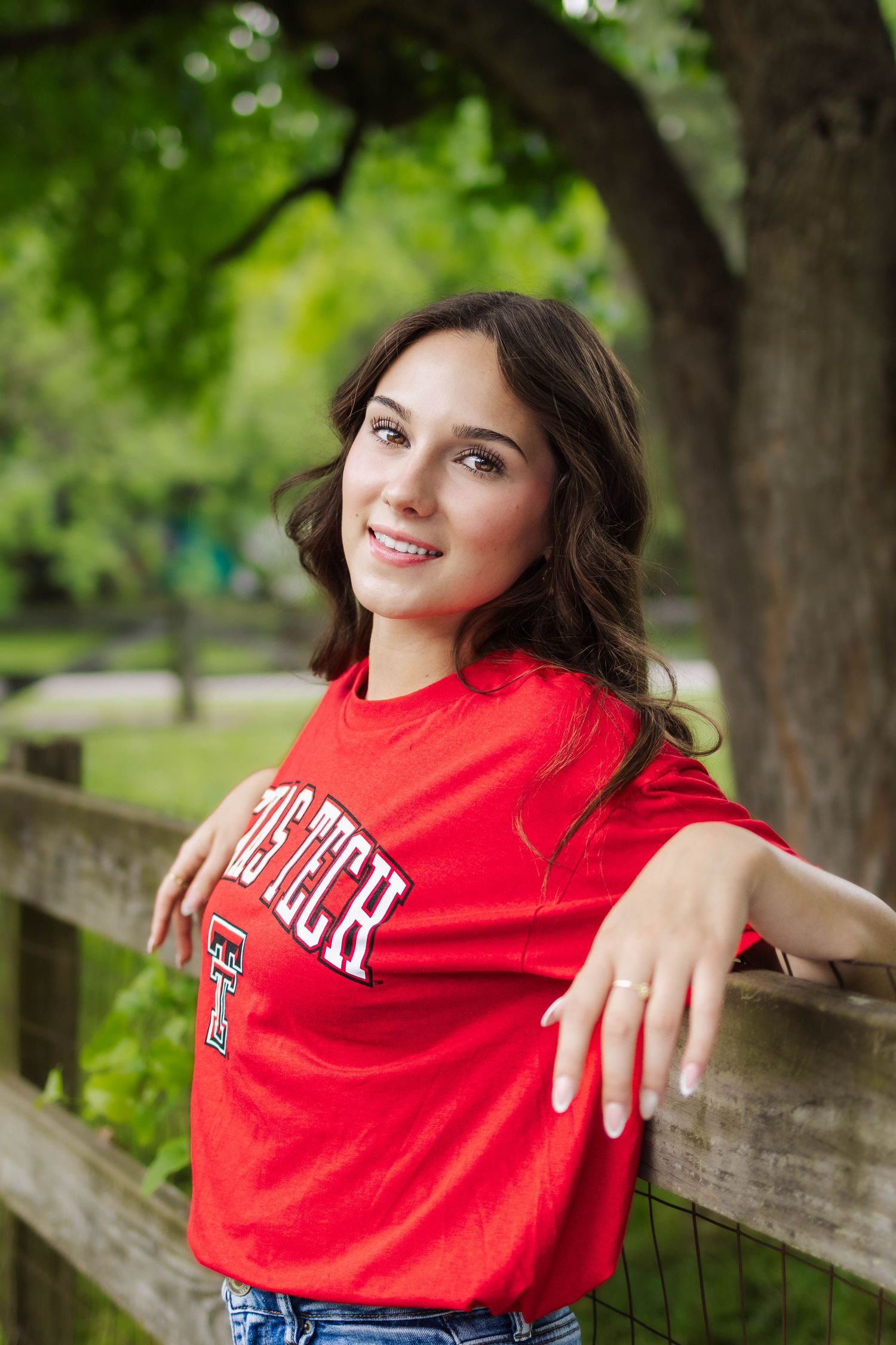 Smiling brunette in red Texas Tech shirt leaning on wooden fence outdoors.